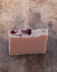 The handcrafted, all-natural Rose Geranium Soap bar sits on a wooden table, with dried rose petals sprinkled on top of the soaps roughly cut edges.
