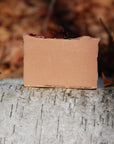 The cold-process soap bar, Rose Geranium, sits on a Silver Birch trunk in a forest in the fall. It's vibrant pink colour from the French Pink Clay highlighted against the white bark of the tree.