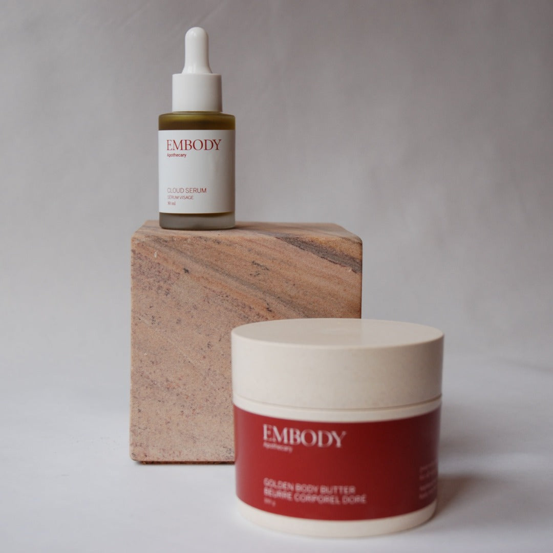 The Cloud Serum, made for sensitive skin, sits on a marble block behind a jar of Golden Body Butter. These products together help to soothe, hydrate, and heal the skin.