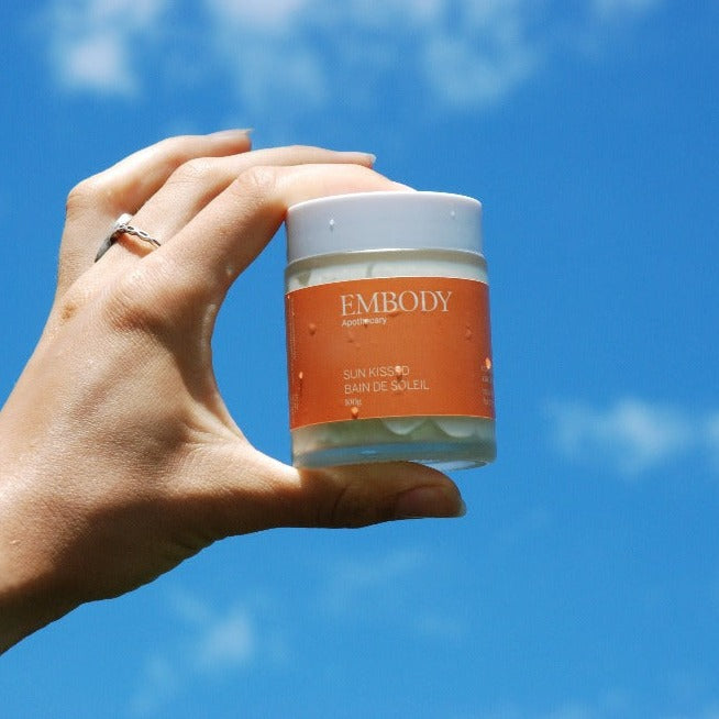 100g jar of Sun Kissed all-natural sunscreen is being held agains the blue sky