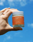 100g jar of Sun Kissed all-natural sunscreen is being held agains the blue sky
