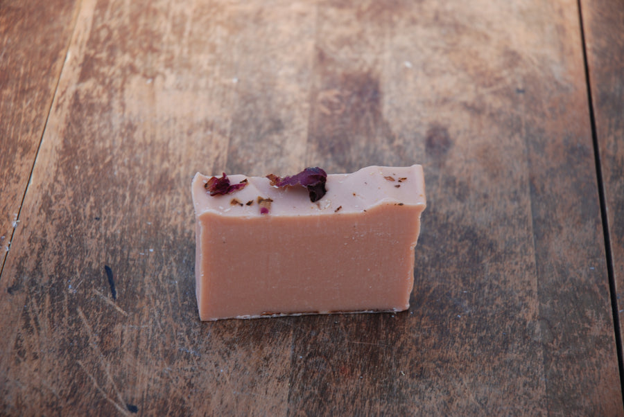 The handcrafted, all-natural Rose Geranium Soap bar sits on a wooden table, with dried rose petals sprinkled on top of the soaps roughly cut edges.