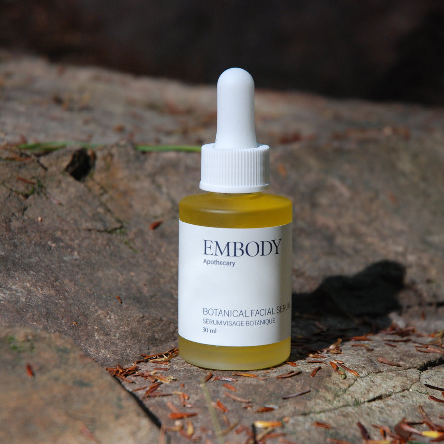 The Botanical Facial Serum sits on a rock, its vibrant colour from its star ingredients, Calendula and Rose Geranium, shining in a ray of sun.